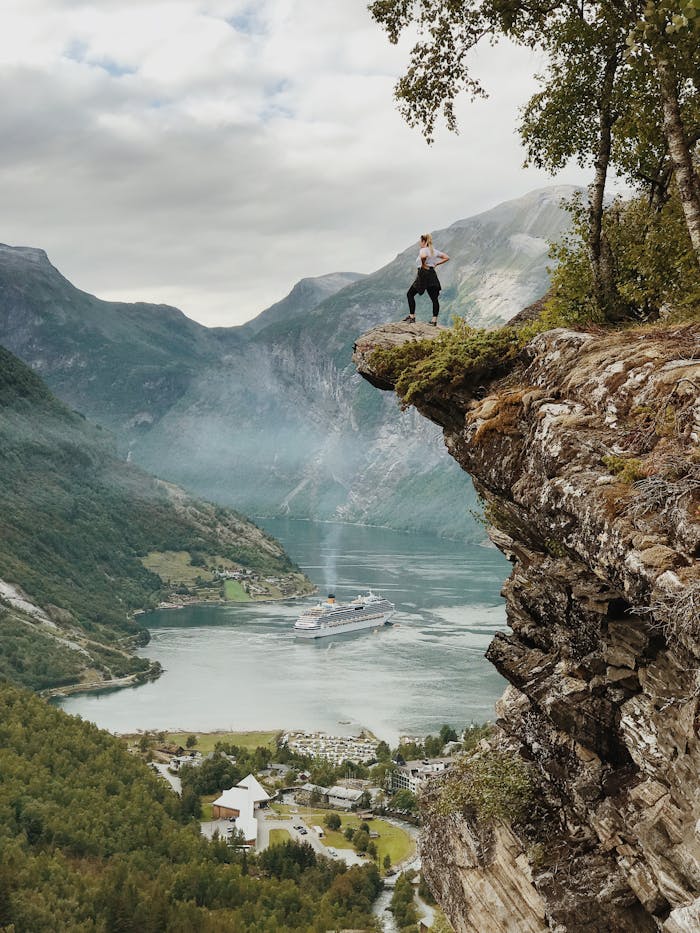 gallery-05 Person enjoys breathtaking view from a cliff above Geiranger Fjord, Norway, with clear skies and cruise ship below.