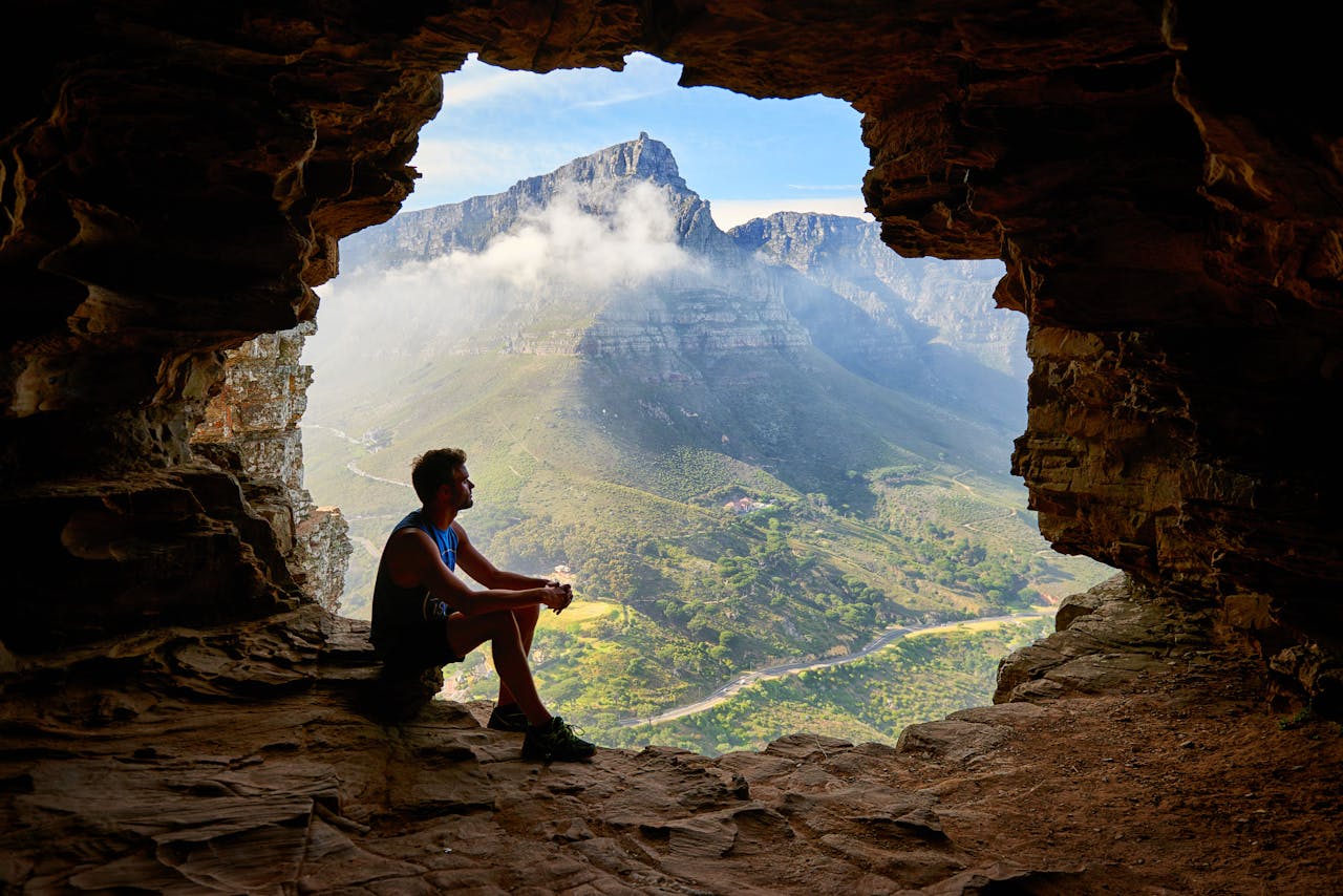 about-img-01 A man sitting in a cave overlooking a majestic mountain landscape under daylight.
