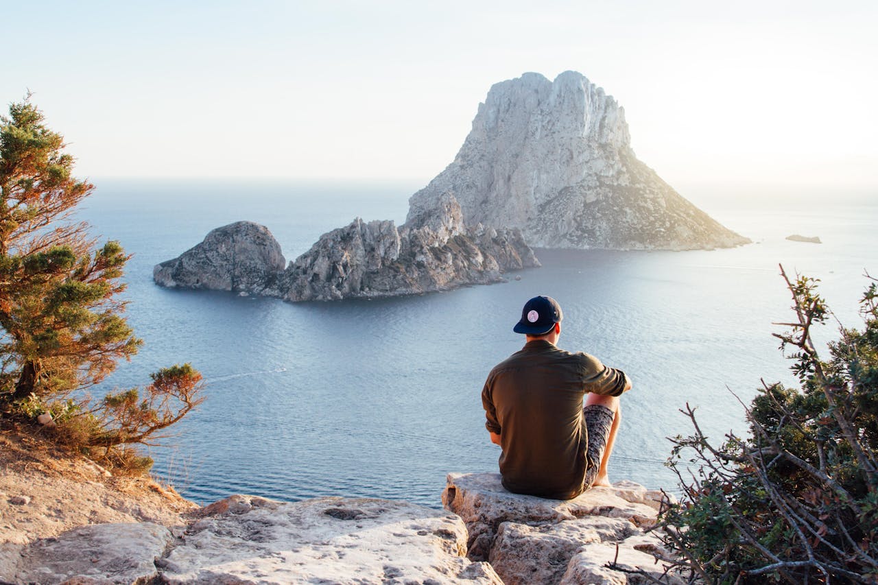 home-img Man enjoys a scenic view of Es Vedrà at sunset from a cliff in San Juan Bautista, providing a perfect summer escape.
